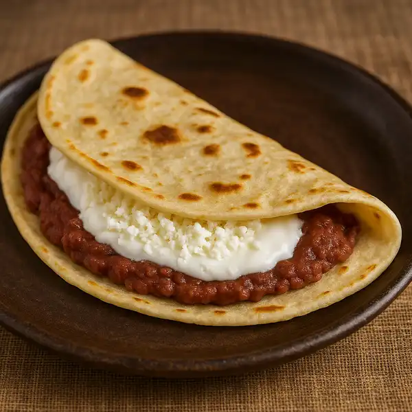 A thick, soft, and lightly toasted flour tortilla is folded in half. The filling is visible at the seam, showing a layer of dark, mashed red beans, a smear of white cream, and a generous sprinkle of crumbled white cheese.