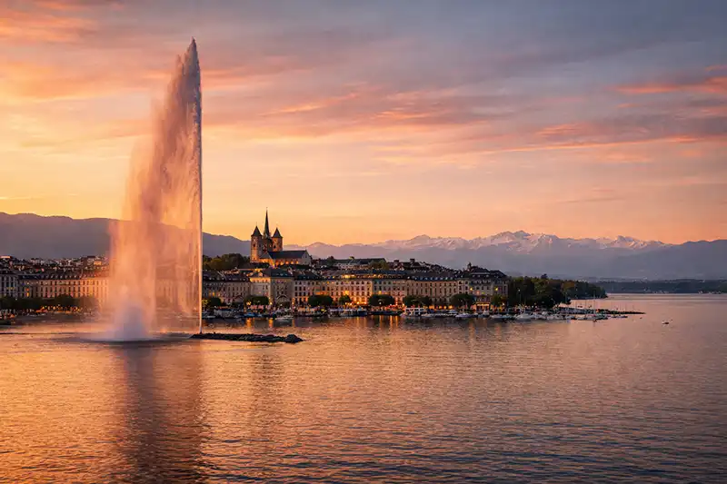 View of the Jet d'Eau fountain on Lake Geneva with the Geneva city skyline and Alps in the background.