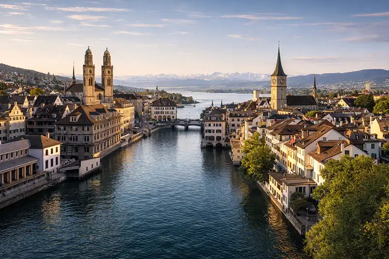 Scenic panoramic view of Zurich Old Town, the Limmat River, Grossmünster church, and distant Swiss Alps.