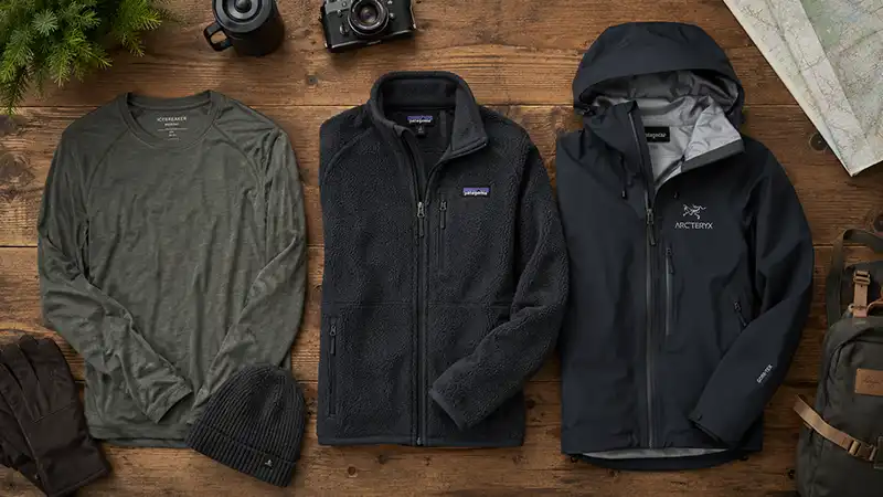 A flat lay of layered cold-weather clothing including a base layer, fleece, and waterproof shell